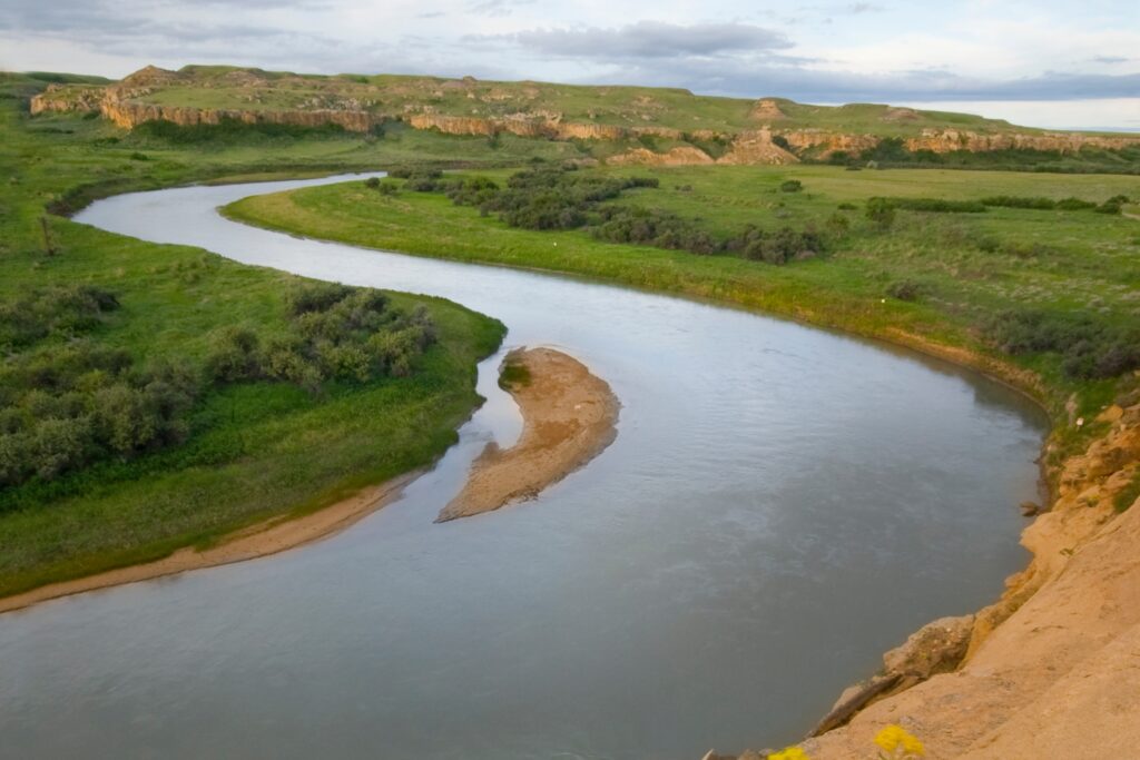 A winding river surrounded by lush green grass and shrubs, with sandbanks along the river’s edge and a backdrop of rolling hills under a partly cloudy sky. //