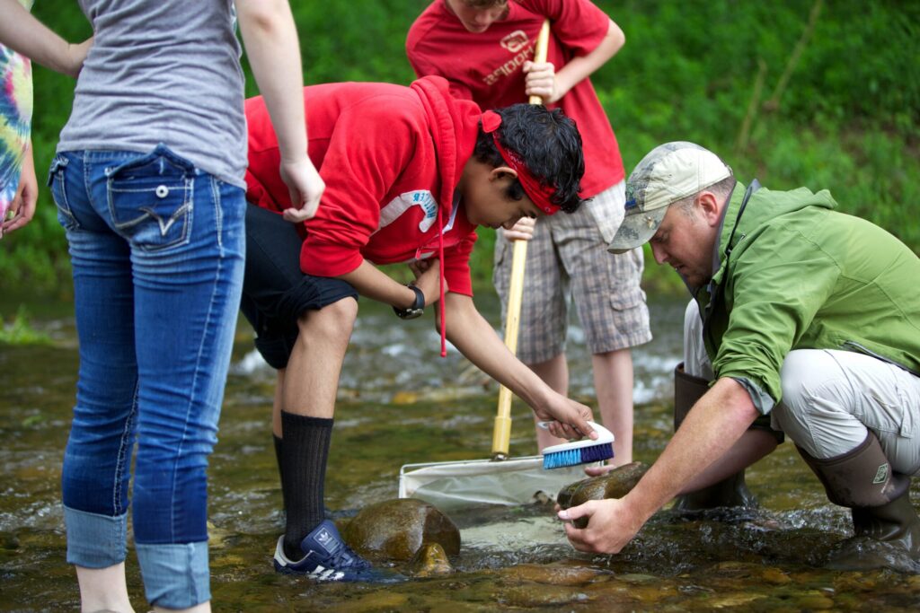 Texte alternatif : un groupe de personnes vêtues de vêtements décontractés se tiennent dans un ruisseau peu profond. Une personne utilise une brosse sur un rocher tandis quune autre tient un filet. La scène se déroule dans un décor naturel avec un feuillage vert à proximité. / Un groupe de personnes en tenue décontractée se tient dans un ruisseau peu profond. Une personne utilise une brosse sur un rocher tandis quune autre tient un filet. La scène se déroule dans un cadre naturel avec de la verdure à proximité.